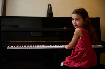 Authentic beautiful little child girl in elegant red dress, looking at camera, sitting at piano and...