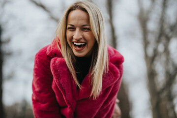 A joyful young blonde woman wearing a striking pink coat is laughing heartily in an outdoor, winter...