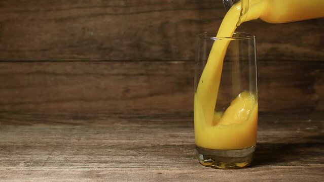 Medium, low shot, glass jug pouring orange juice into a glass, brown rustic wooden setting, copy space to the left