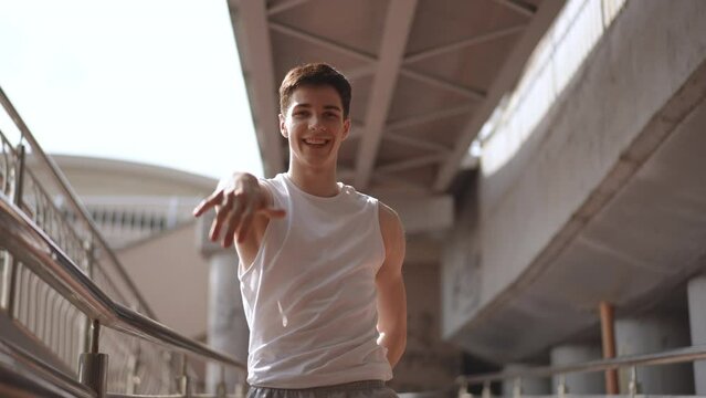 Low Angle Shot. Happy young guy energetically moves his body and arms while standing under a city bridge outdoors. The guy dances, smiles and looks at the camera