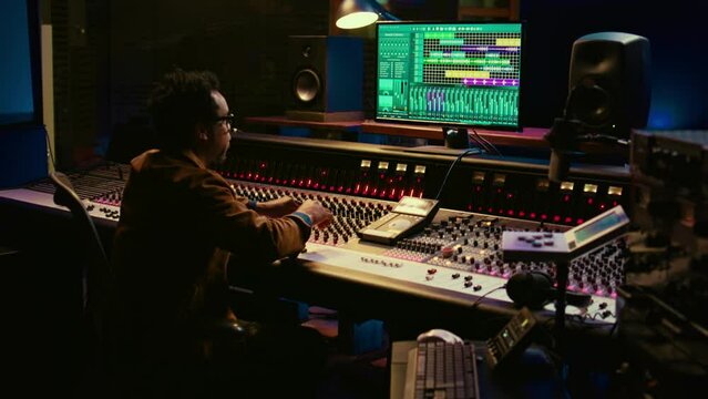 African american sound expert operates on audio console with moving faders and colored meters, editing in professional studio control room. Producer works on album, recording hit song. Camera B.
