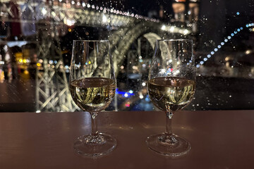Looking at Porto Luiz I bridge through the window in the rainy night, with two glasses of porto blanc wine - 771831973