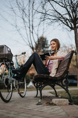 A relaxed young woman sits on a park bench with her legs stretched, reading a book, with a bicycle beside her.