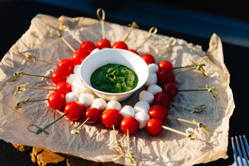 Caprese Skewers with Fresh Basil Pesto Dip. A delightful arrangement of Caprese skewers, featuring cherry tomatoes, mozzarella balls, and a vibrant basil pesto dip on natural paper.