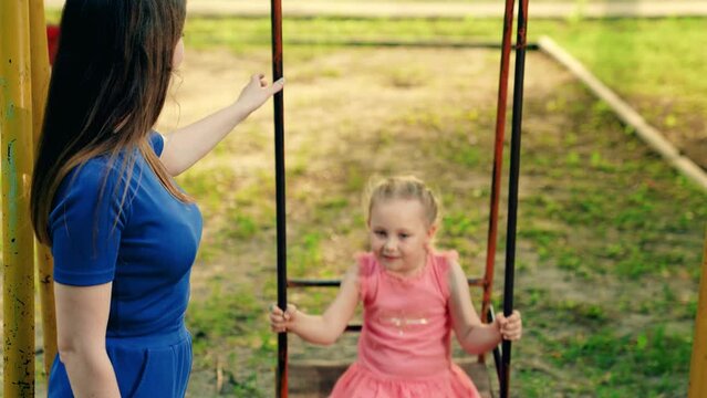 Child has fun on playground in outdoor. Happy family kid mama. Parent mom playing with daughter, city park. Child plays on swing on summer playground. Mother shakes children swing, happy Girl smiling.