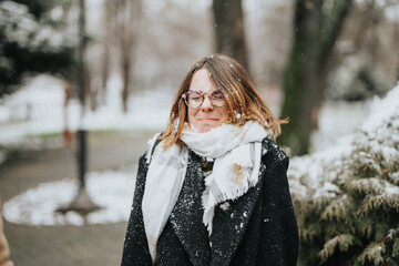 A young businesswoman in a coat and scarf smiles outdoors during a light snowfall, capturing a moment of winter joy in a city park.