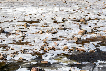 A shallow river covered with shards of broken ice supported by mud and rock. Some water flows among...