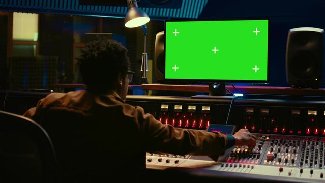 African american audio engineer uses greenscreen on pc in professional recording studio, working with mixing console and twisting knobs to edit tracks. Music producer creating tunes. Camera B.