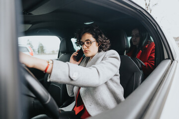 Two professional women in business attire are in a car one is focused on driving while the other is busy with a phone call.