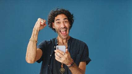 Surprised man with curly hair, dressed in blue shirt, reading good news on mobile phone celebrates...