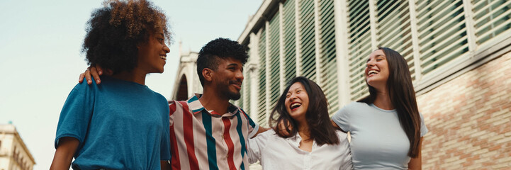 Happy multiethnic young people walk embracing on summer day outdoors, Panorama. Group of friends are talking and laughing merrily while walking along on the street - obrazy, fototapety, plakaty