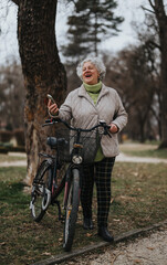 Happy mature woman enjoying a break with her bike in a serene park setting, smart phone in hand.