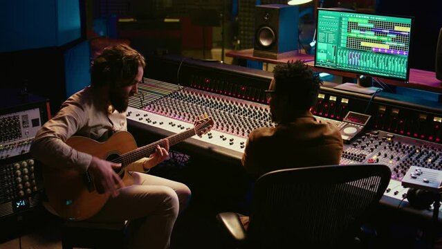 Rockstar recording music on his guitar in professional studio, creating new music for his album in control room. Artist composer producing tracks on electro acoustic guitar. Camera B.