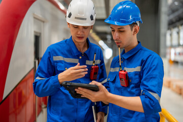 Two engineers in hard hats and blue uniforms are discussing train maintenance details on a tablet...