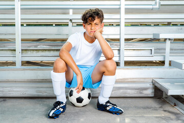 Youth male soccer player sitting on a soccer ball waiting for his chance to play the game