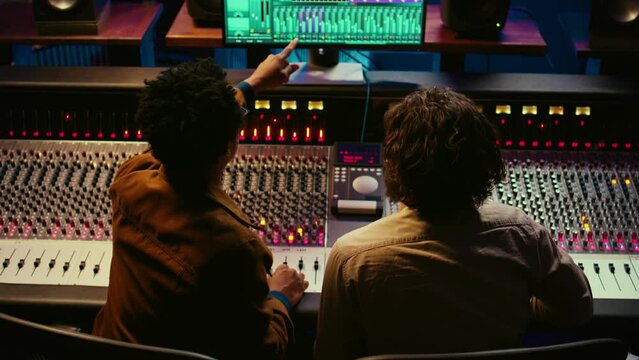 Team of sound engineers working on recording and editing tracks in professional studio control room, collaborating on mix and master audio files. Musician and technician pressing sliders. Camera B.