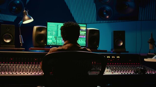 African american music producer vibing on his new track in professional studio, mixing and mastering tunes in control room. Technician pressing buttons and sliders on soundboard panel. Camera B.