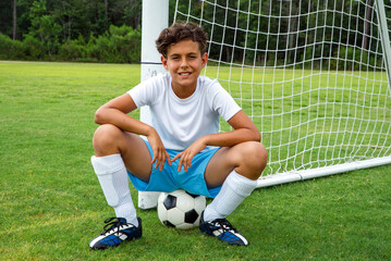 Smiling young male soccer player sitting on a soccer ball in the goal, looking happy after winning a game
