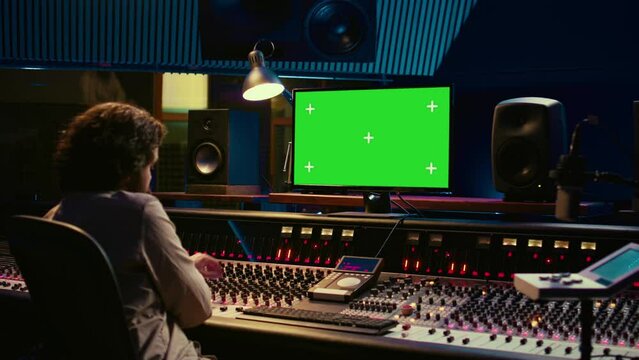 Sound designer editing and recording tracks with mixing console in control room, twisting knobs and pressing key buttons. Audio engineer producing music with greenscreen on monitor. Camera B.