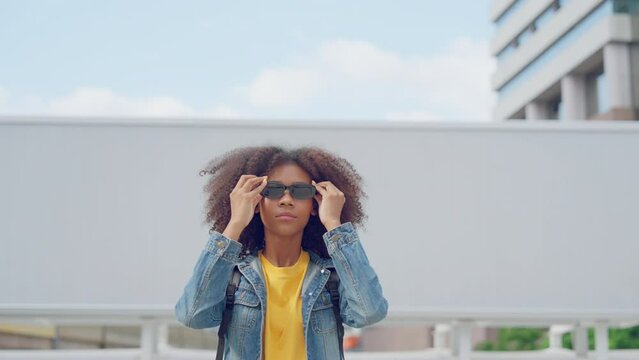 Happy young afro woman walking and wearing sunglasses while wearing yellow shirt and jeans jacket outdoor looking at camera
