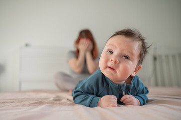 A three-month-old boy lies on his stomach on the bed and his mother sits behind him and cries....