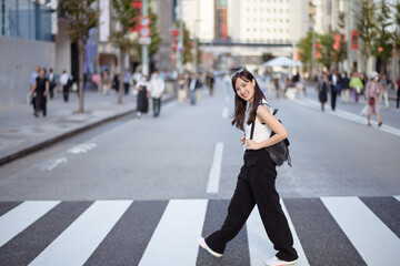 Urban joy unfolds as a young Asian woman navigates a busy crosswalk, seamlessly blending into the lively rhythm of local life. - obrazy, fototapety, plakaty