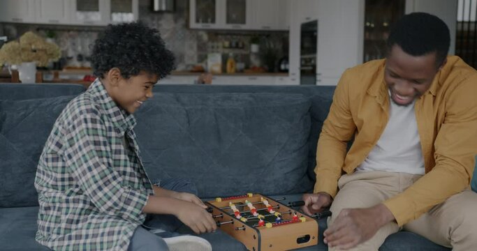 Happy African American dad and son playing foosball having fun in kitchen at home. Table football game and childhood activity concept.
