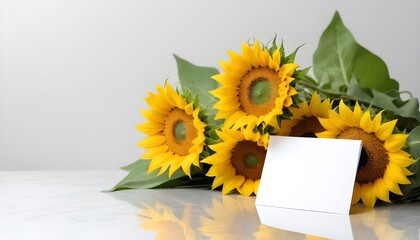 A blank white card in front of a bouquet of yellow sunflowers with green leaves on a reflective white surface