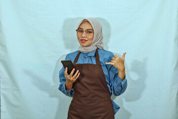 Asian Muslim woman wearing hijab, glasses and brown apron, holding a mobile phone and making phone...