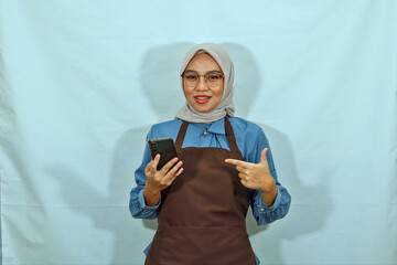young Asian Muslim woman wearing hijab, glasses and apron, pointing at her smartphone with a finger and smiling showing her teeth isolated on a white background. housewife muslim lifestyle concept
