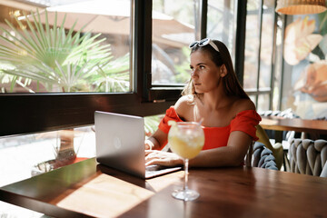 Woman works on laptop in a sunny cafe, glass of juice nearby. The setting is modern, with natural light illuminating the space. The image captures a moment of focused work.