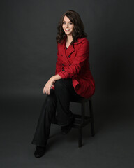 full length portrait of beautiful brunette woman model, wearing red trench coat jacket with black leather pants. sitting pose with hand gesture on dark grey studio background.