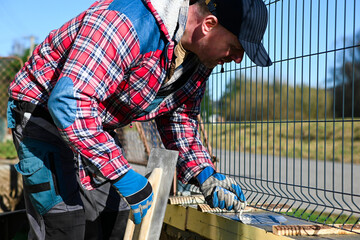 A man in overalls and gloves is repairing the fence in front of the family house. Close-up view and blurred background