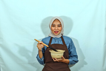 beautiful Asian woman in a brown apron and hijab holding a spoon and bowl while doing housework. smile showing her teeth isolated over a white background. housewive Muslim concept