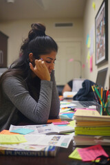 A focused student is engrossed in her studies at a desk littered with study materials and a laptop - obrazy, fototapety, plakaty