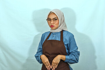 young Asian Muslim woman wearing hijab, glasses and brown apron while looking at camera isolated on white background. People housewife muslim lifestyle concept
