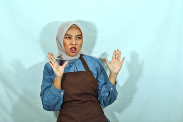 shocked young Asian Muslim woman in hijab and brown apron showing stop gesture with palm refusing...