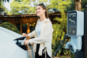 Young woman and sustainable urban commute with EV electric car recharging at outdoor cafe in...
