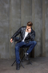 Modern young man with a stylish hairstyle, wearing a cool black leather jacket and sitting on a chair on a dark background