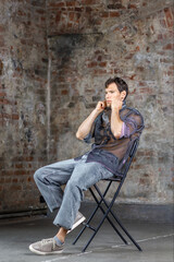 A modern young man with a stylish hairstyle sits on a chair on a dark background.
