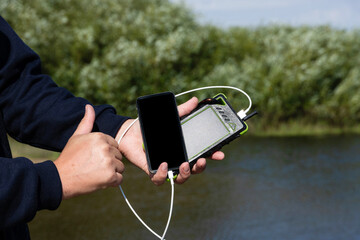 Tourist with smartphone and charger in the forest, on the river in summer. The guy with the powerbank is lost in nature. The concept of modern technology to always stay connected and connected.