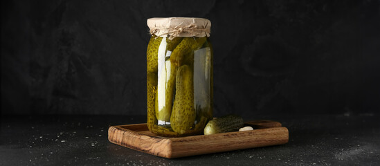 Wooden board with jar of tasty pickled cucumbers and garlic on black background