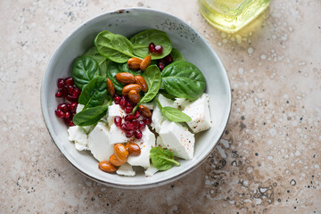 Grey bowl with feta cheese, baby spinach, pomegranate and almond salad, horizontal shot on a beige granite background, elevated view