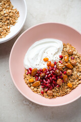 Greek yogurt with granola, pomegranate and nuts in a roseate bowl, vertical shot on a light-beige stone surface, selective focus