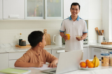 Smiling father bringing glass of juice and porridge to son doing homework at kitchen counter