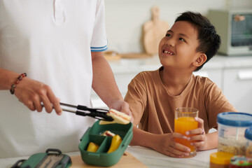 Smiling Vietnamese boy drinking orange juice when father packing him lunch box