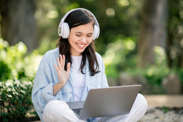 Engaging young woman with headphones, waving at her laptop screen
