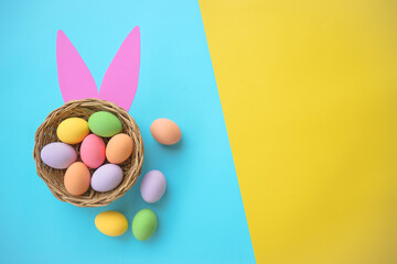 Fototapeta na wymiar Top view of colorful Easter eggs in basket with pink bunny ears on a blue and yellow two-tone background with copy space happy easter