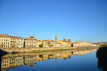 Fototapeta na wymiar A distant view of traditional Italian houses by the riverbank of Arno River in Pisa against a cloudless sky. The background texture features village homes in Europe with water reflection