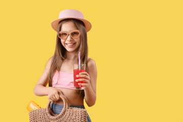 Cute little girl in swimsuit, sunglasses with wicker bag and glass of juice on yellow background